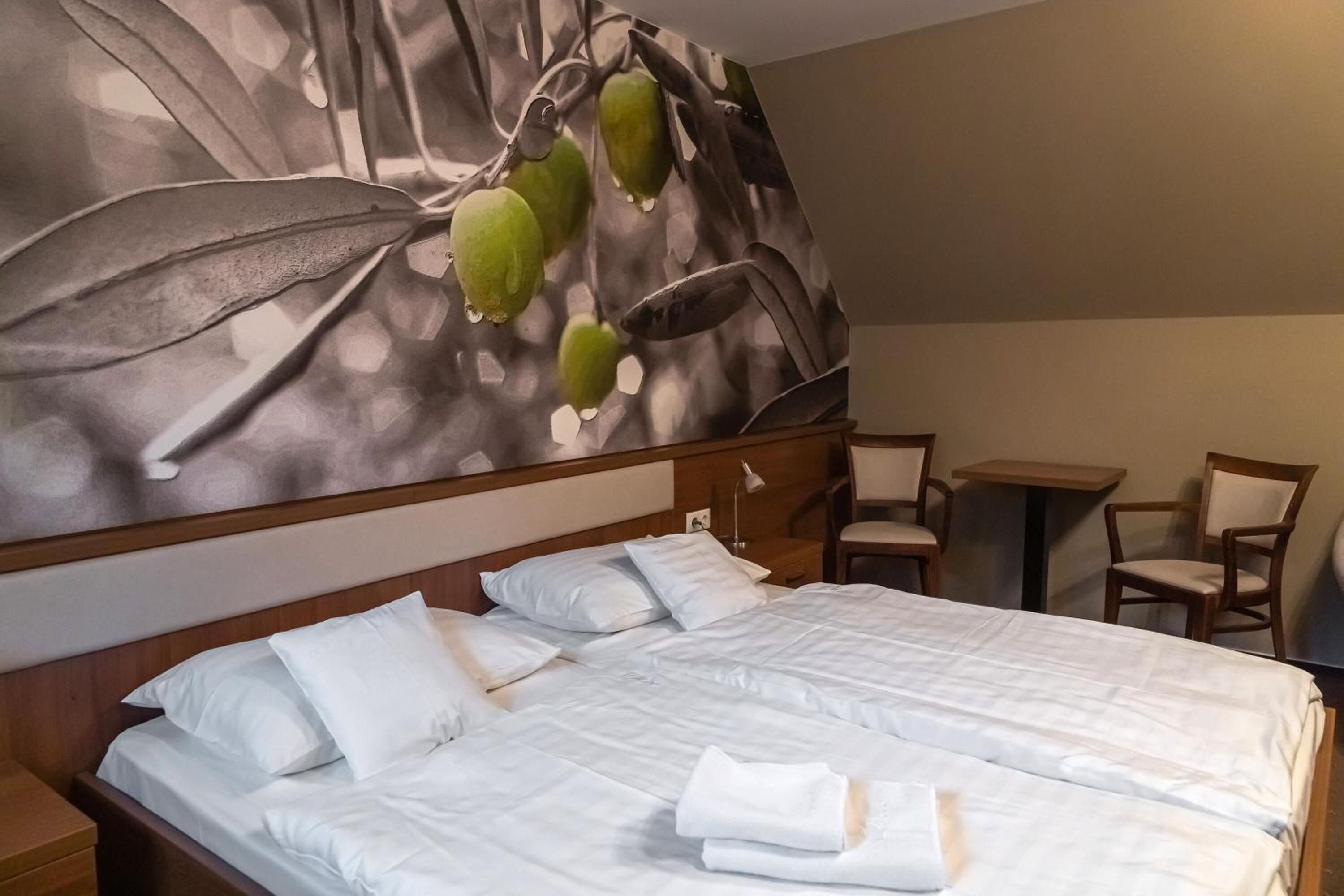 A hotel room with a large bed, a wooden headboard, and a wall mural of leaves and fruit. There are two chairs and a small table.