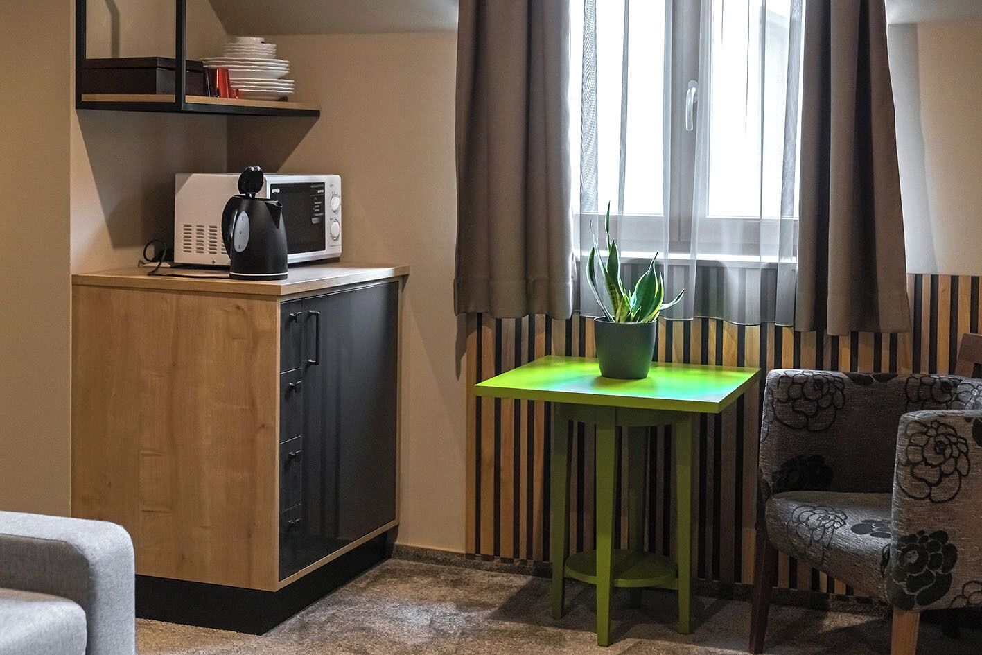 A compact room with a kitchenette, a green table with a potted plant, and a patterned chair.