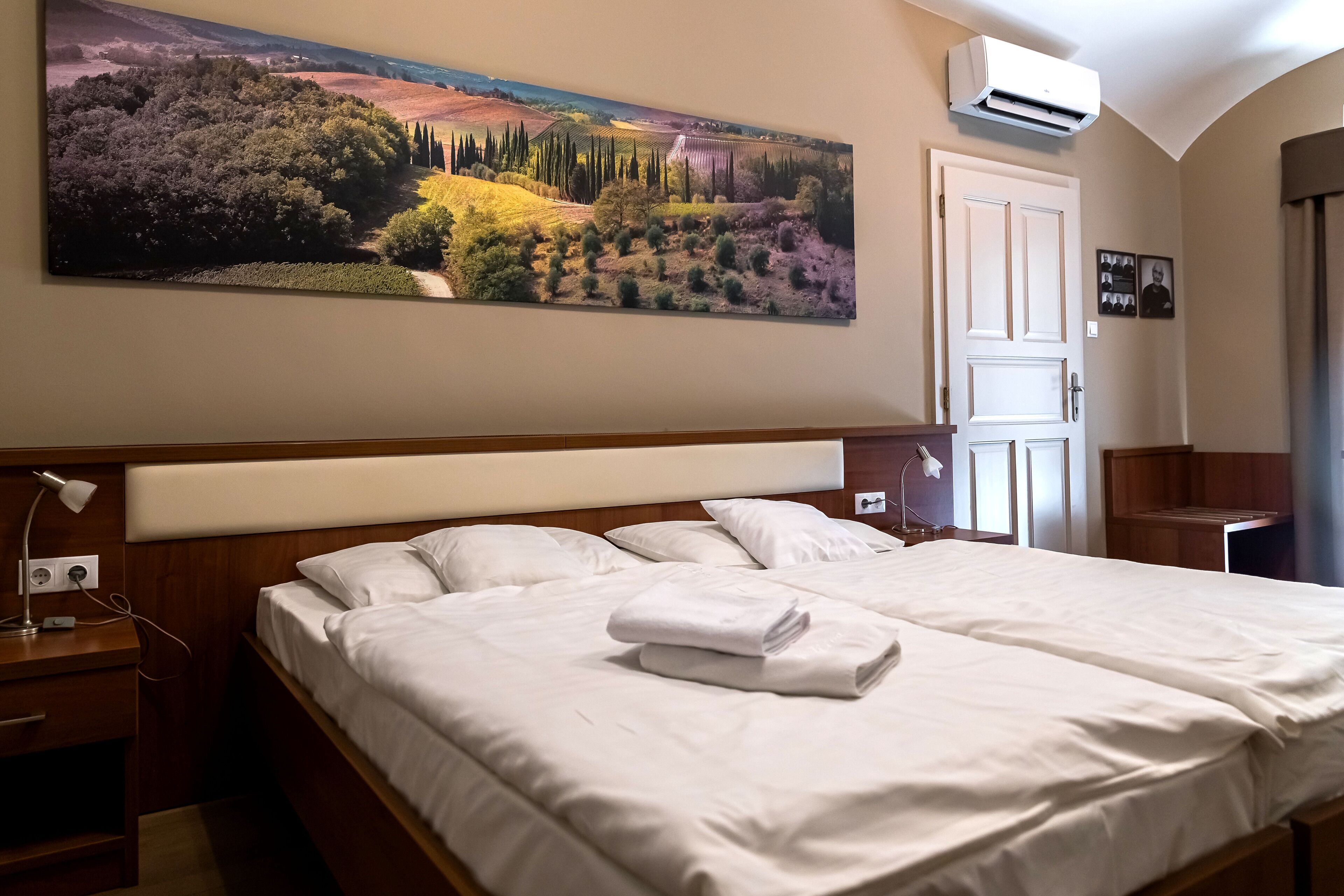 A hotel room with a large landscape painting, a double bed, a bedside table with a lamp, and an air conditioning unit on the wall.