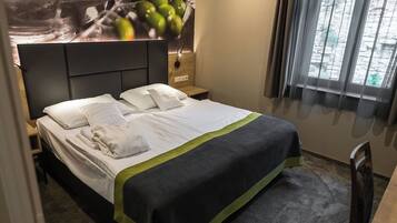 A neatly made bed with white linens and a green accent, a nightstand with a lamp, and a window with curtains.