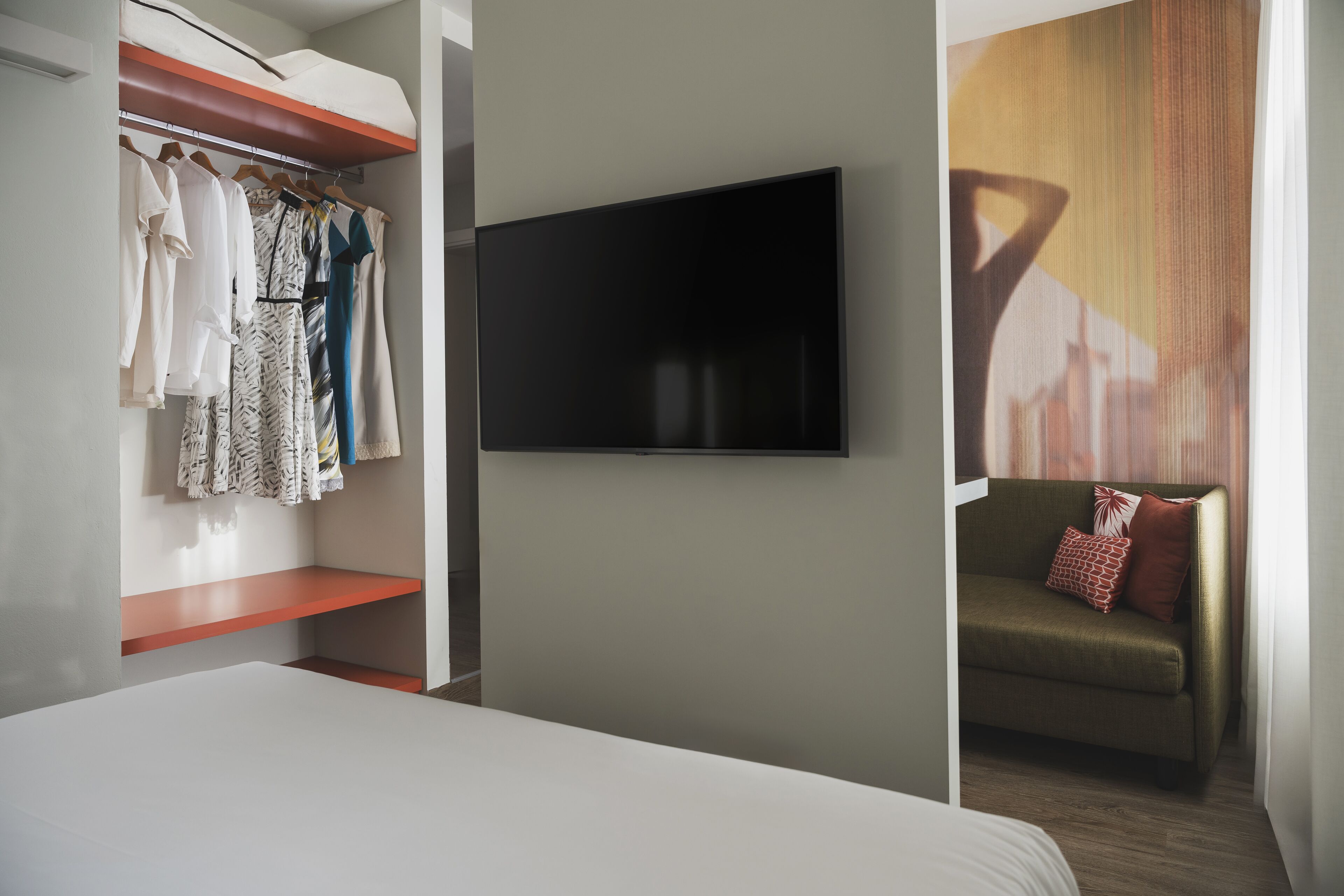 Habitación de hotel con cama, televisor montado en la pared, armario con ropa colgada y sofá con cojines.