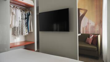Habitación de hotel con cama, televisor montado en la pared, armario con ropa colgada y sofá con cojines.