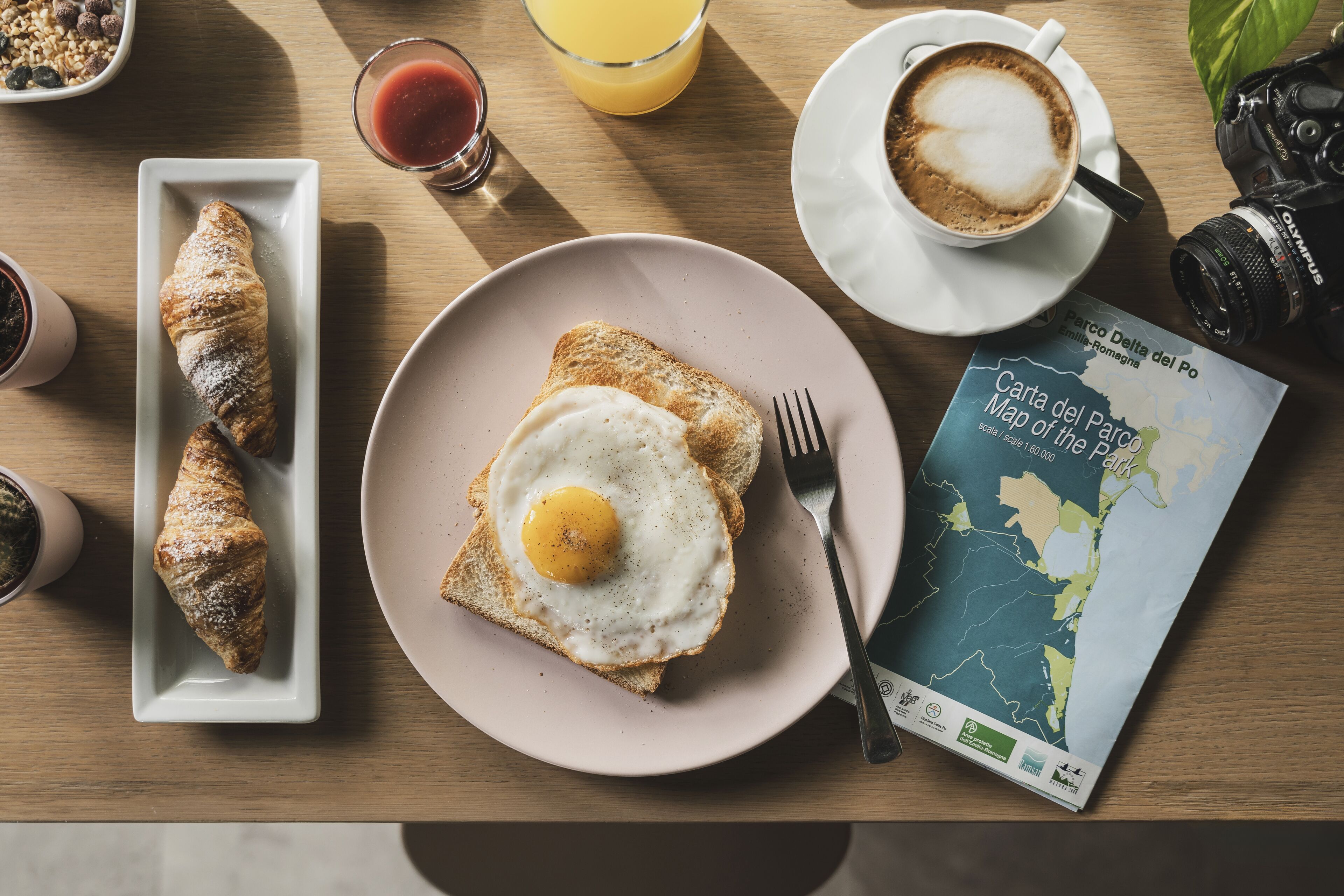 Un desayuno con cruasanes, tostadas con huevo, café, un mapa y una cámara sobre una mesa de madera.