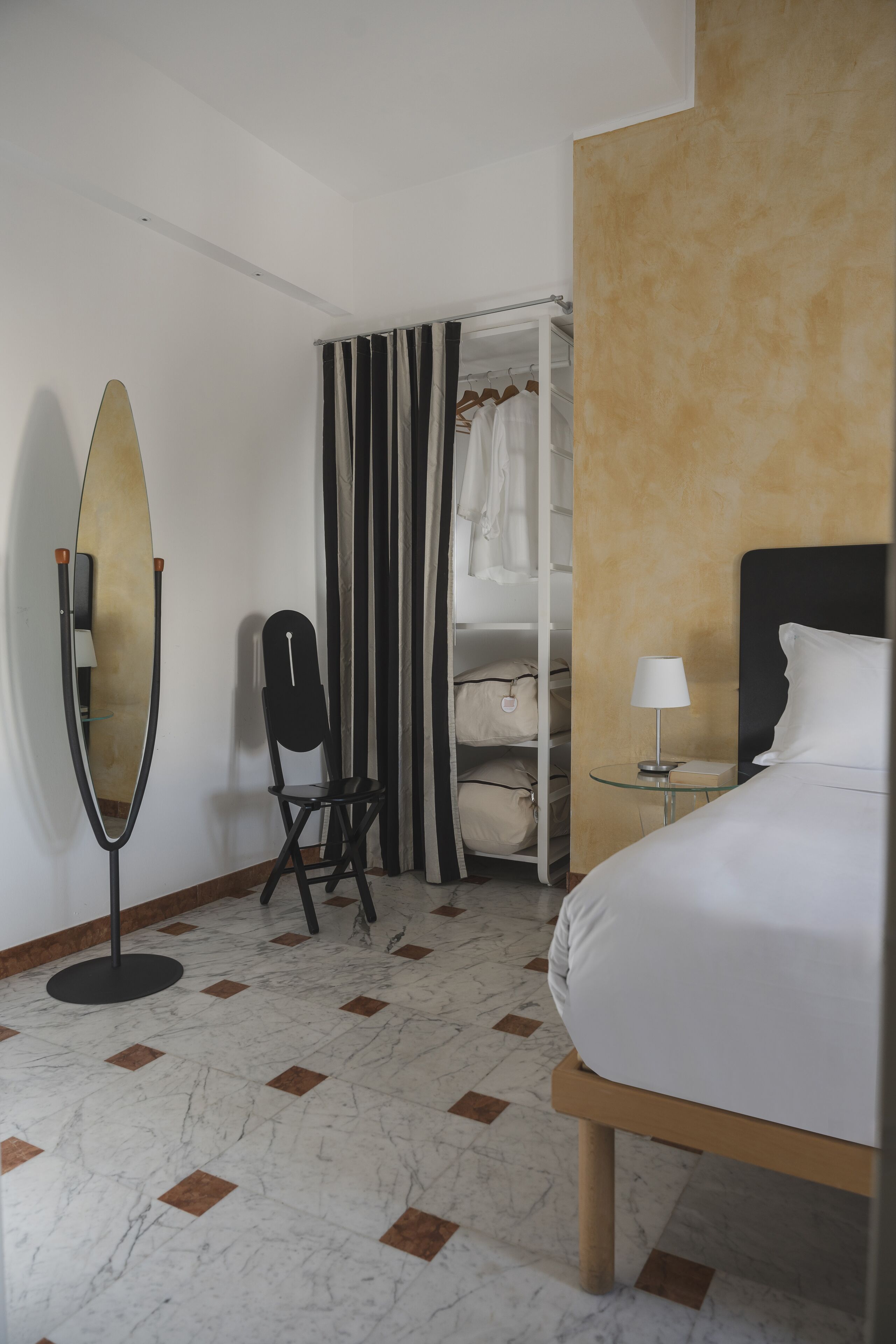 Une chambre d’hôtel comprenant un lit, une table de chevet avec une lampe, une chaise, un porte-manteau et un miroir.