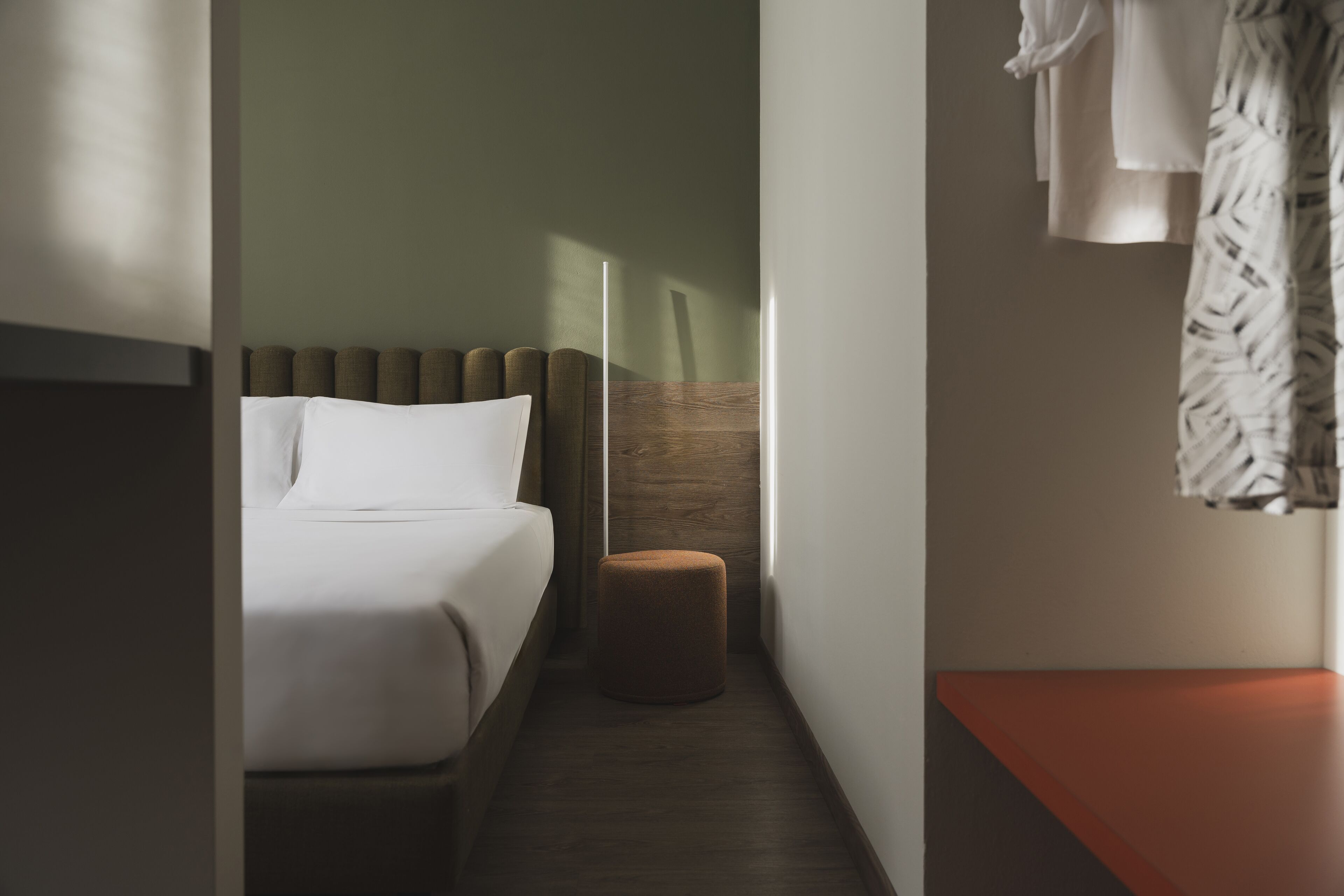 A narrow hotel room with a bed, a small stool, and a bench.