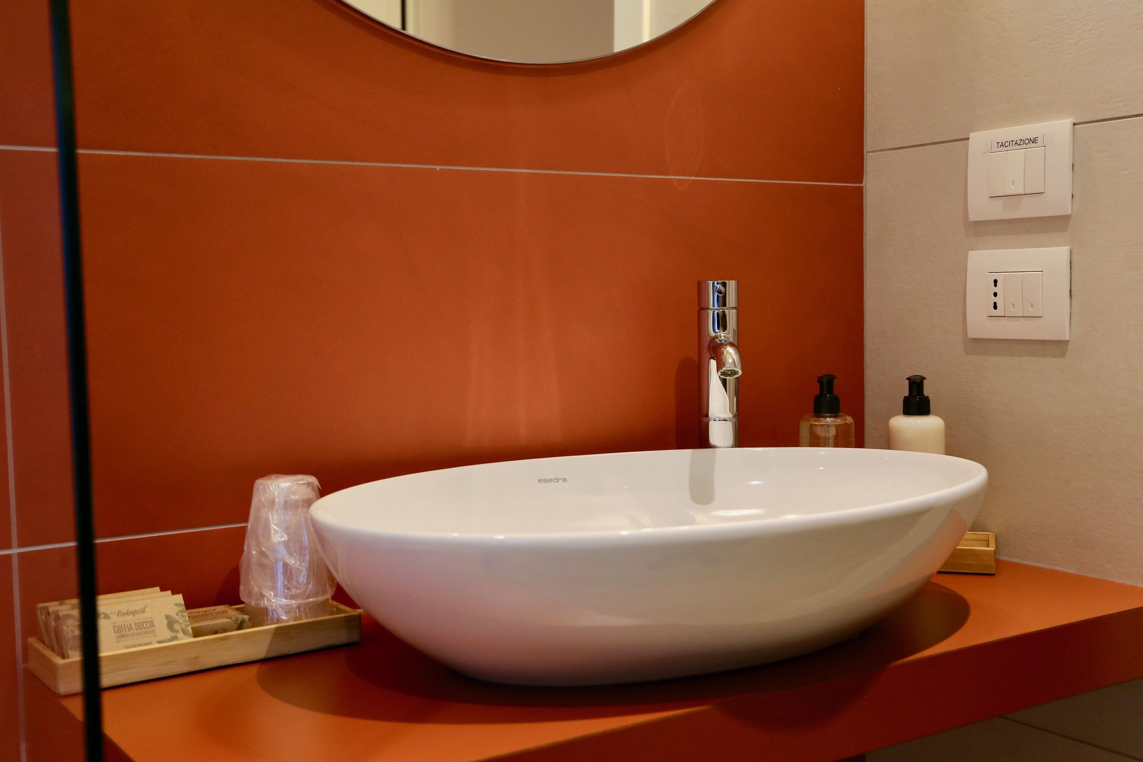 A modern bathroom with a large white sink, a chrome faucet, and a mirror above it. There are toiletries on the counter and a wooden tray with items on it.