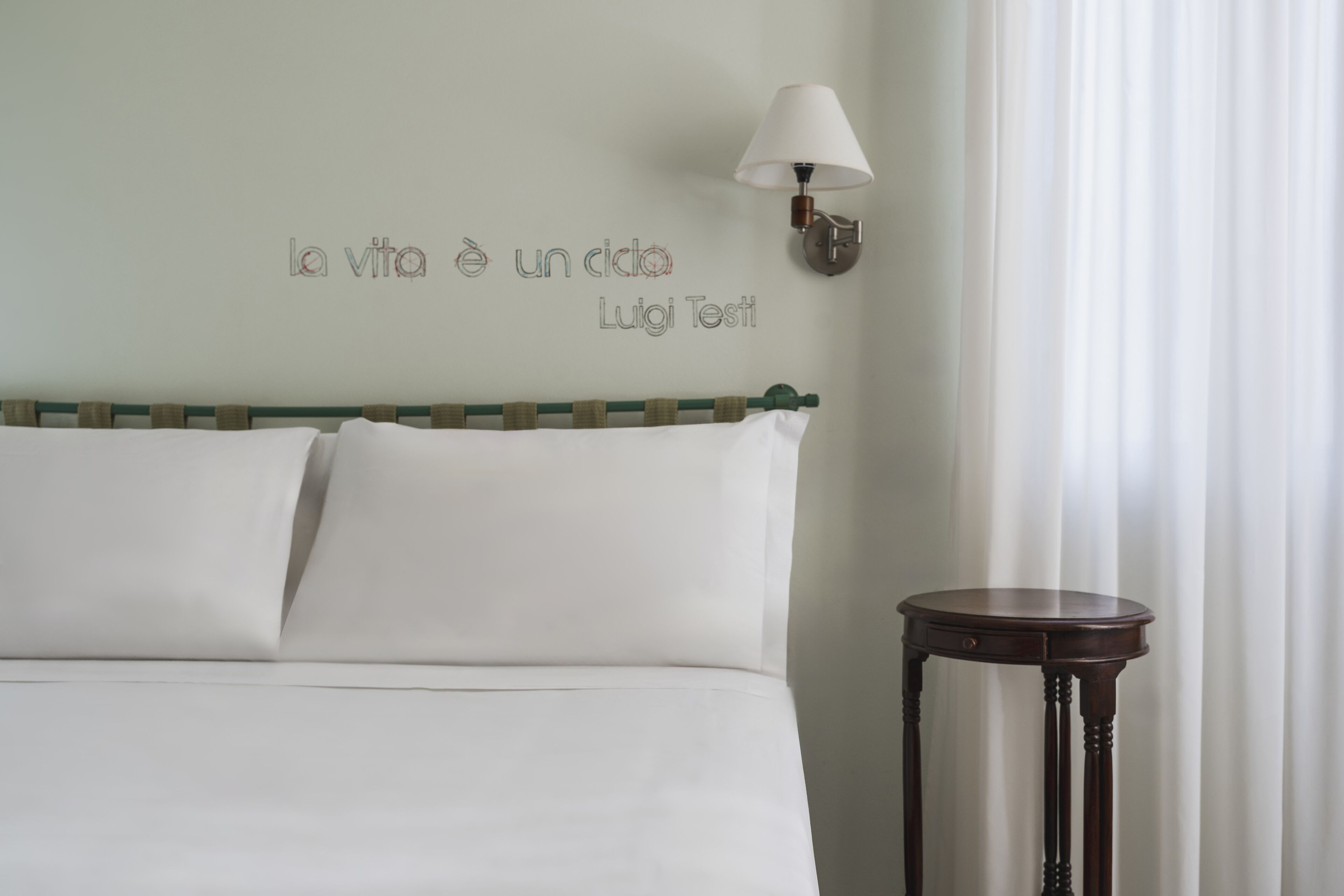 Una cama bien tendida con sábanas blancas, una lámpara de pared y una mesita de noche de madera.