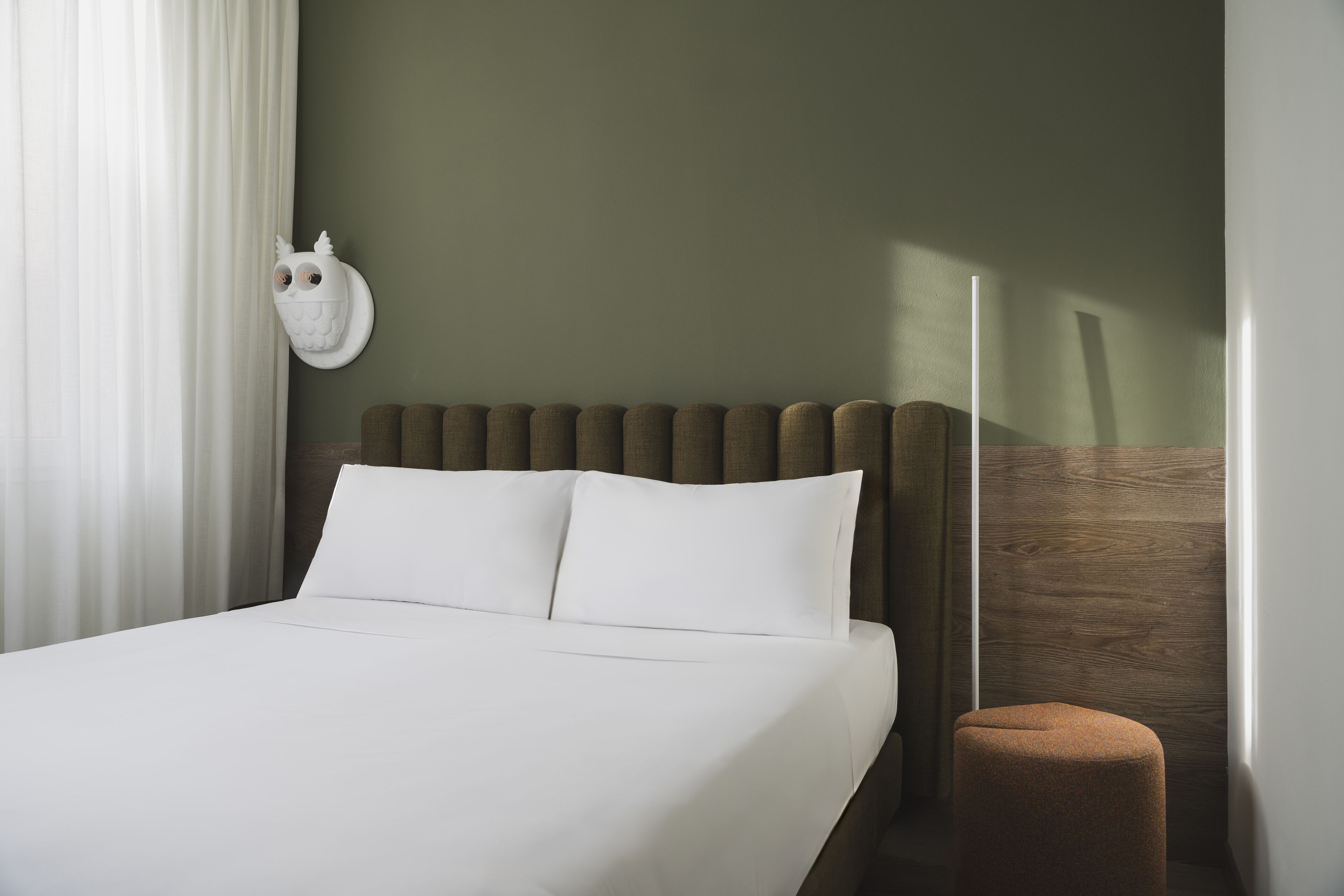 A neatly made bed with white linens, a headboard with vertical slats, and a small brown ottoman beside it.