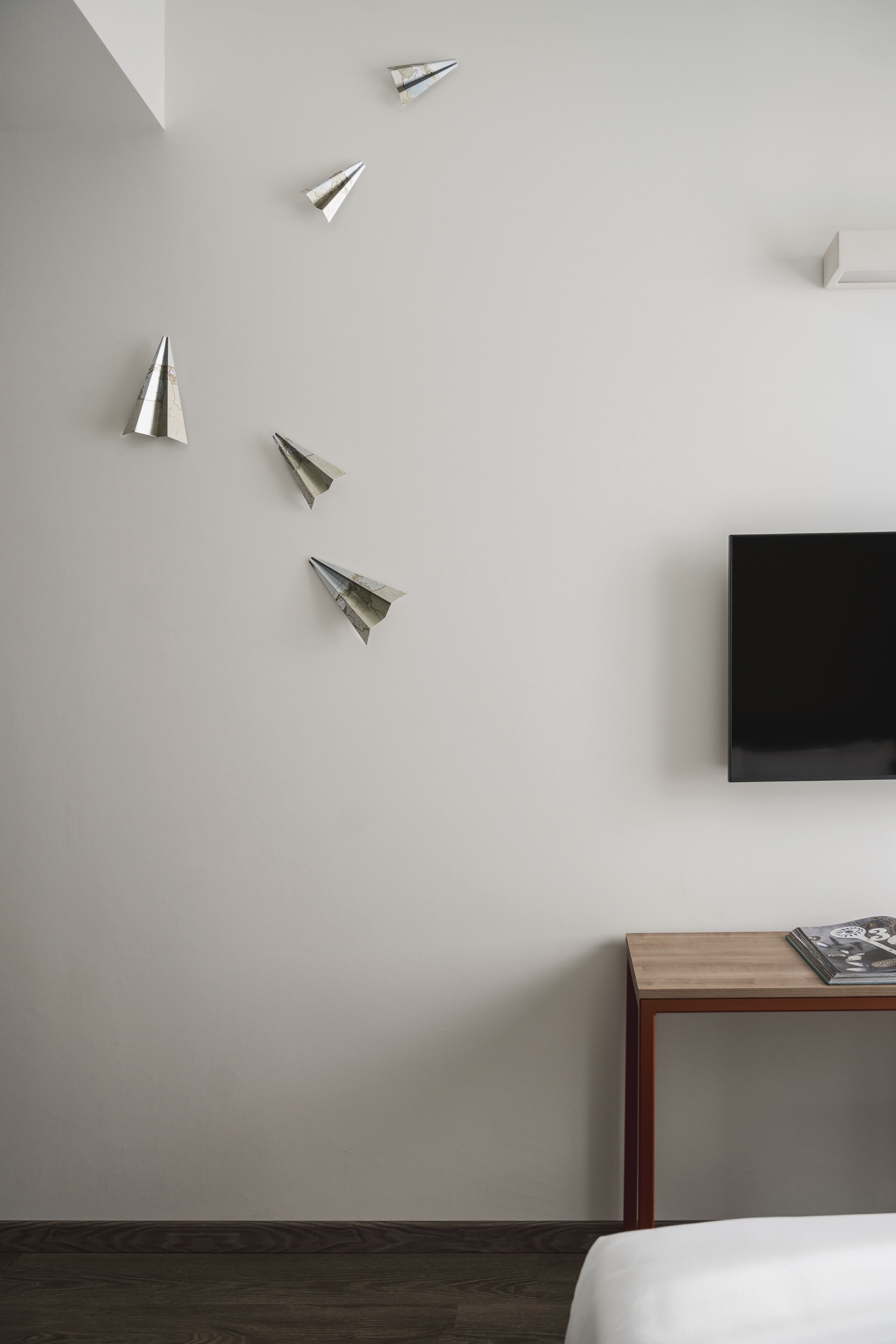 A modern hotel room with a bed, a wooden nightstand, a flat-screen TV, and four paper airplanes on the wall.