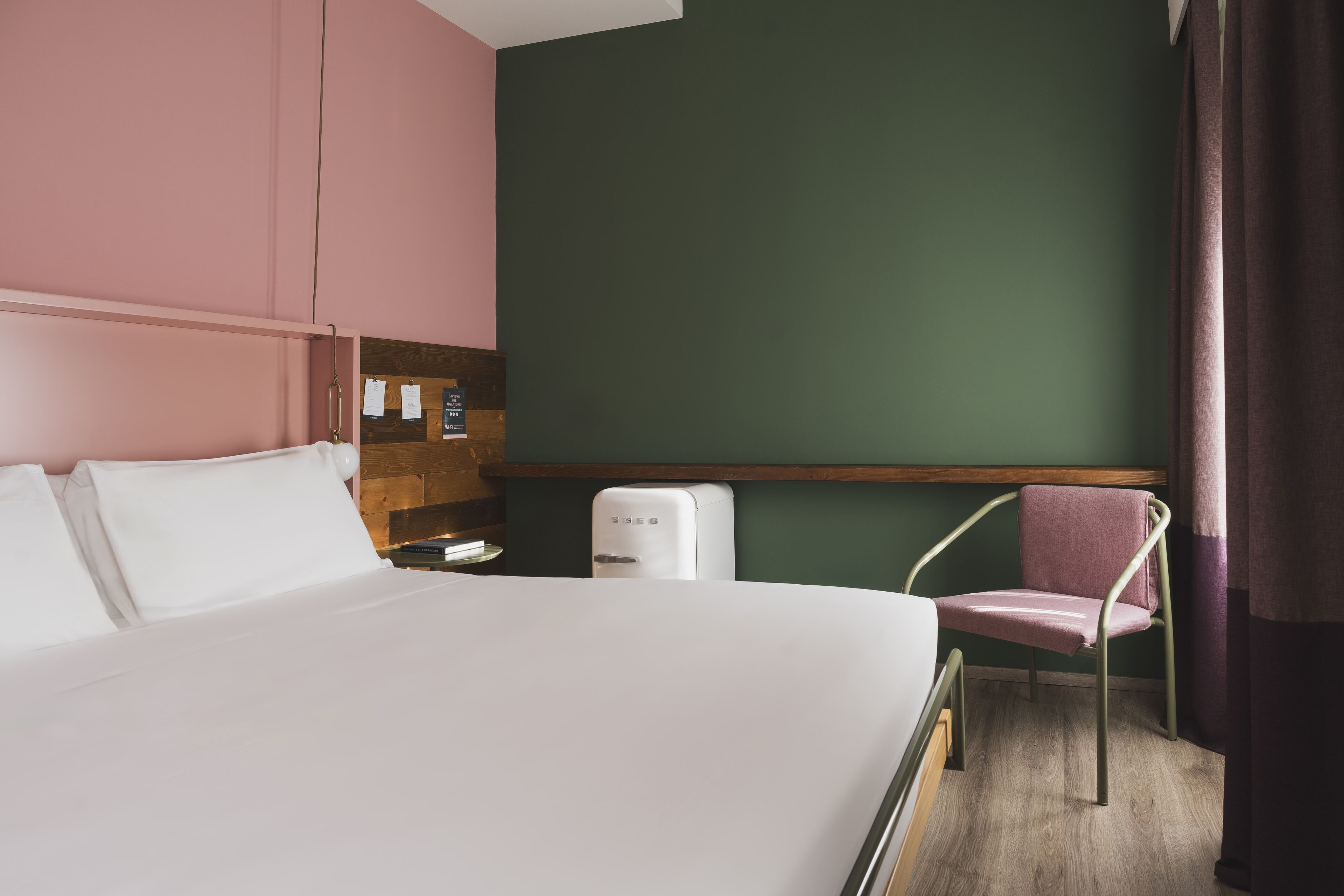 Habitación de hotel con cama, una silla, una mesita pequeña, estante en la pared y un reloj de pared.
