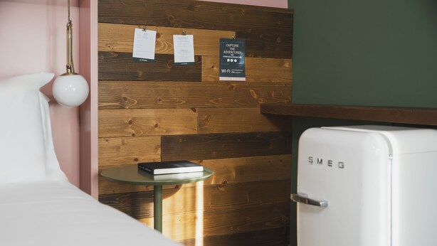 A room with a wooden wall, a white bed, a small table with a book, a white refrigerator, and a wall-mounted sign. - Al Ponticello Room & Breakfast (Comacchio)