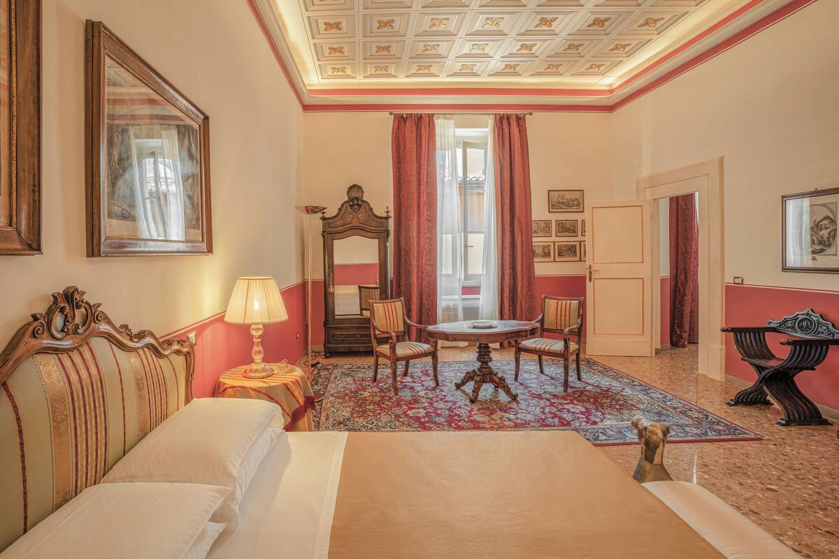Une chambre d’hôtel comprenant un lit, une table avec des chaises, un bureau, une lampe, un miroir et une fenêtre avec des rideaux.