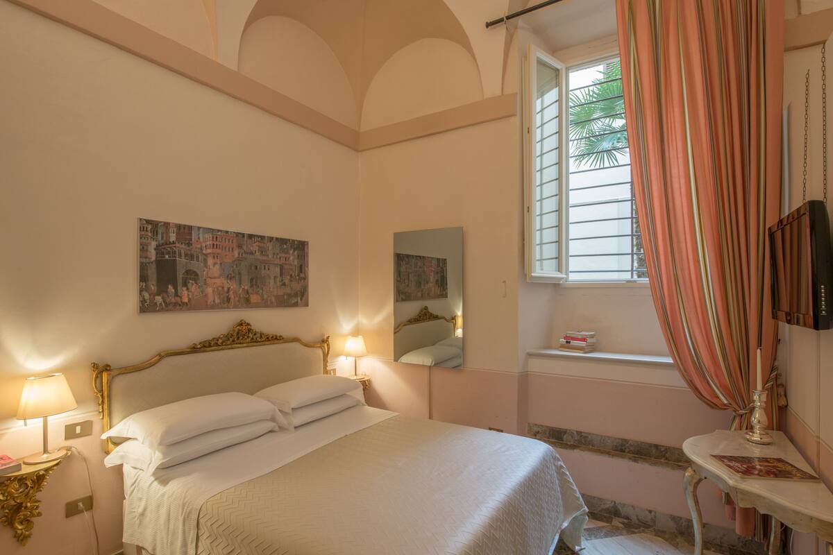 Une chambre à coucher avec un lit, des lampes de chevet, une fenêtre avec des rideaux, un miroir et des œuvres d’art accrochées au mur.