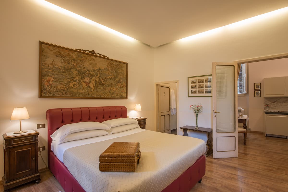 Une chambre d’hôtel avec un grand lit, deux tables de chevet, un tableau accroché au mur, une porte donnant sur une salle de bain et une petite table sur laquelle est posé un vase.