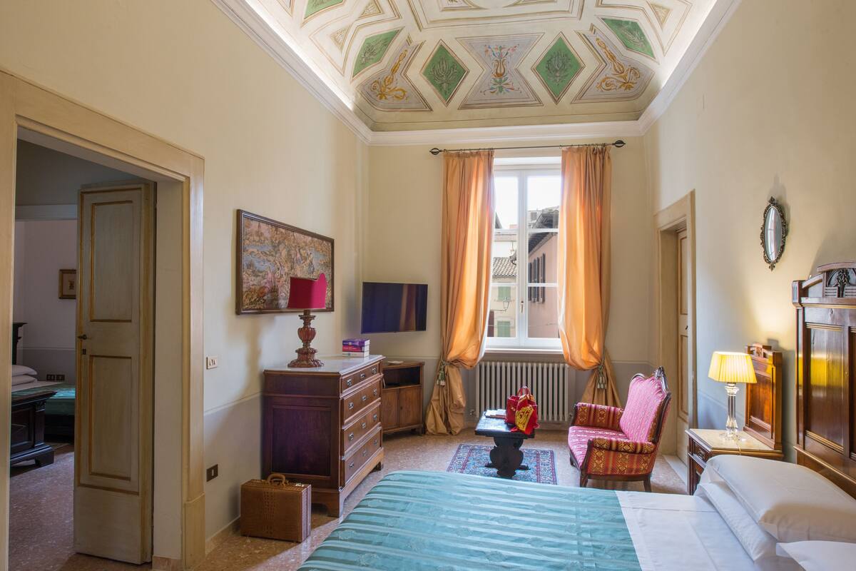 Une chambre d’hôtel comprenant un lit, un bureau, une chaise, une commode, une fenêtre avec des rideaux et un plafond orné de motifs géométriques.