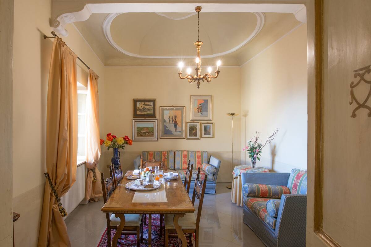 Un coin repas avec une table en bois, des chaises et un lustre. On y trouve également un canapé agrémenté de coussins colorés et un vase rempli de fleurs.
