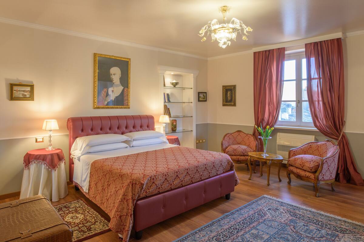 Une chambre à coucher avec un grand lit, un lustre, un coin salon avec des fauteuils et une fenêtre avec des rideaux.