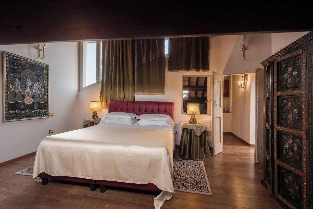 Une chambre à coucher avec un grand lit, un plancher en bois, une table de chevet, une lampe, une porte et un mur orné d’un tableau encadré.