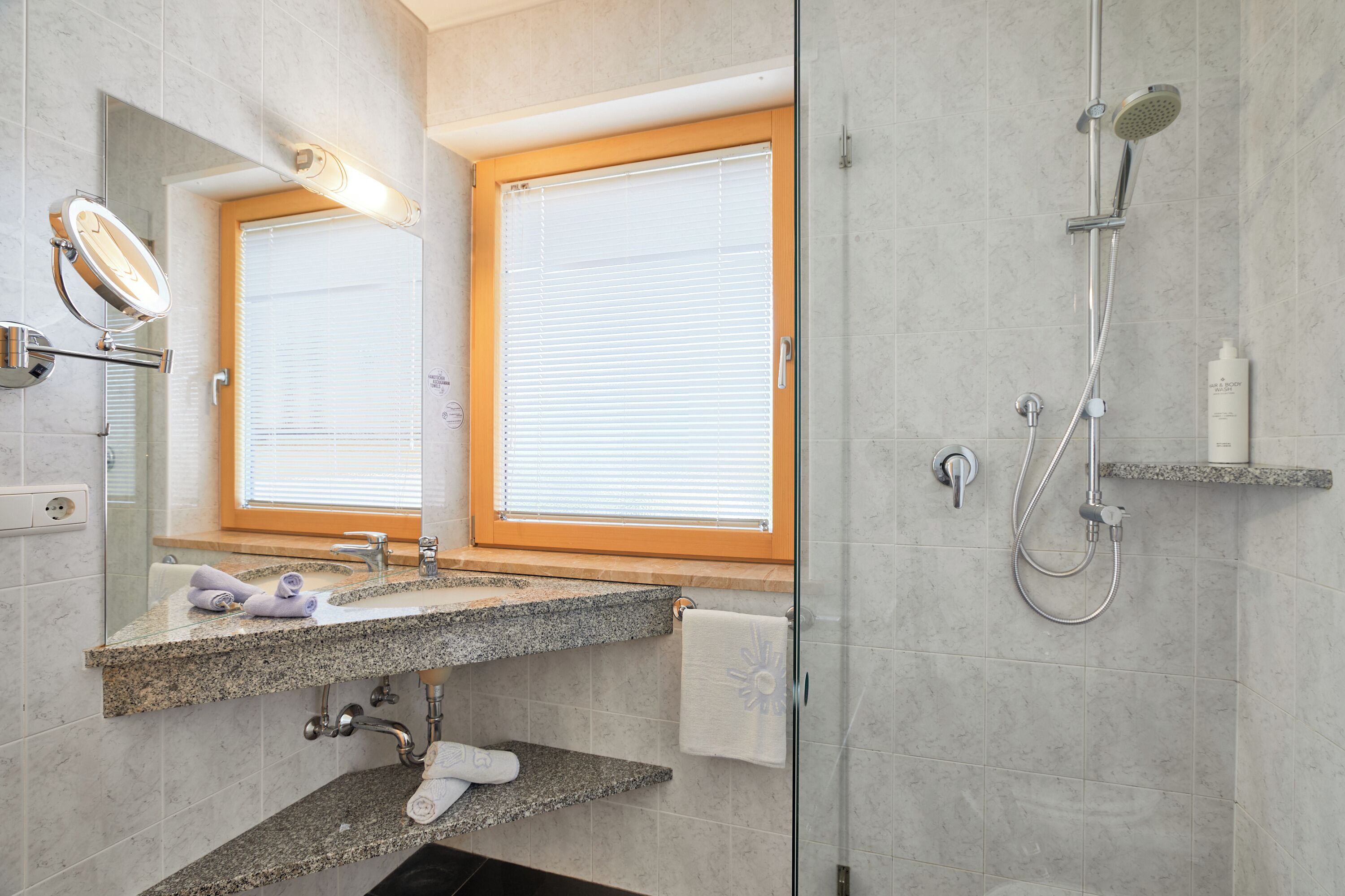 A bathroom with a granite countertop, a mirror, a towel rack, a shower with a handheld showerhead, and a window with blinds.