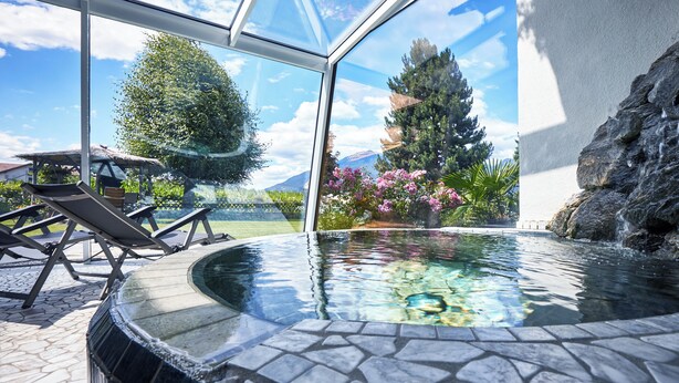Een glazen hottub met een stenen omlijsting, met uitzicht op een tuin met een boom en een waterval.