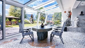 Een oranjerie met een stenen tafel en stoelen, grote glasramen en uitzicht op een tuin.