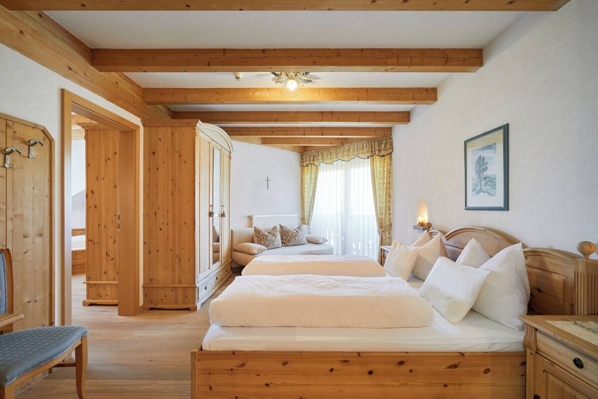 Una habitación con vigas de madera, una cama con ropa blanca, un banco, un armario de madera y un cuadro enmarcado en la pared.