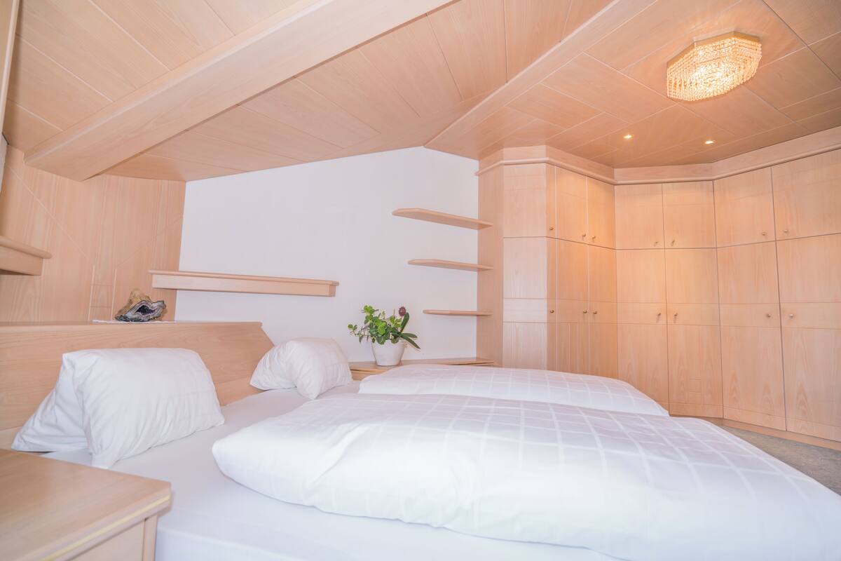 Un dormitorio moderno con techo de madera, una cama grande con ropa blanca, una mesita de noche con una planta y armarios empotrados de madera.
