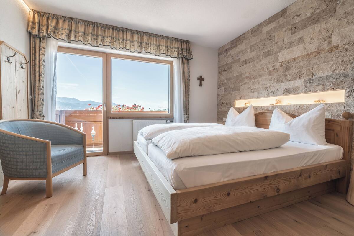 Un dormitorio con una cama de madera, una silla, un ventanal con cortinas y vistas a montañas.