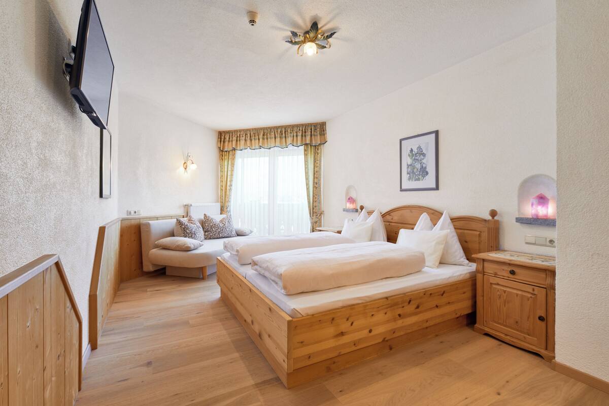 Un dormitorio con cama de madera, mesita de noche, lámpara de cabecera, televisor montado en la pared, ventana con cortinas y una lámpara en el techo.