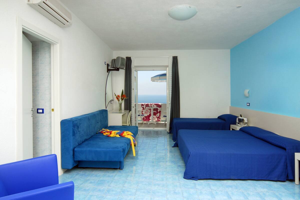 A hotel room with two beds, a sofa, a TV, and a view of the sea.