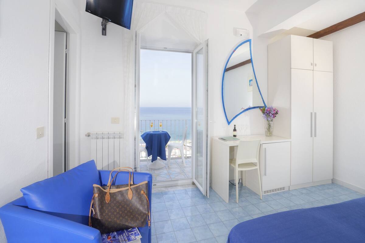 A hotel room with a bed, a blue sofa, a desk with a chair, a wardrobe, a TV mounted on the wall, and a balcony with a view of the sea.