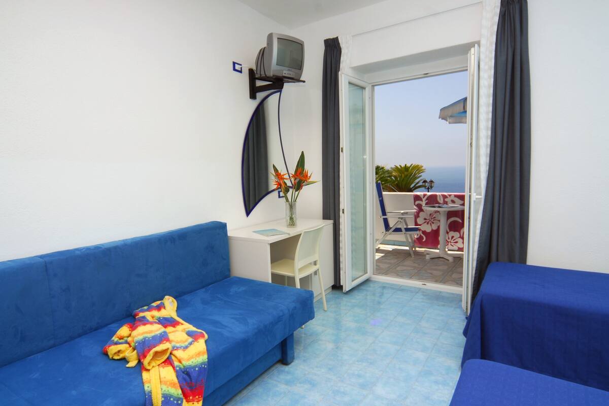 A room with a blue sofa, a small desk with a TV, and a view of the sea through a sliding door.