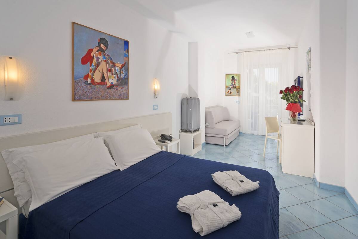 A hotel room with a bed, a sofa, a desk with a vase of flowers, and artwork on the walls.