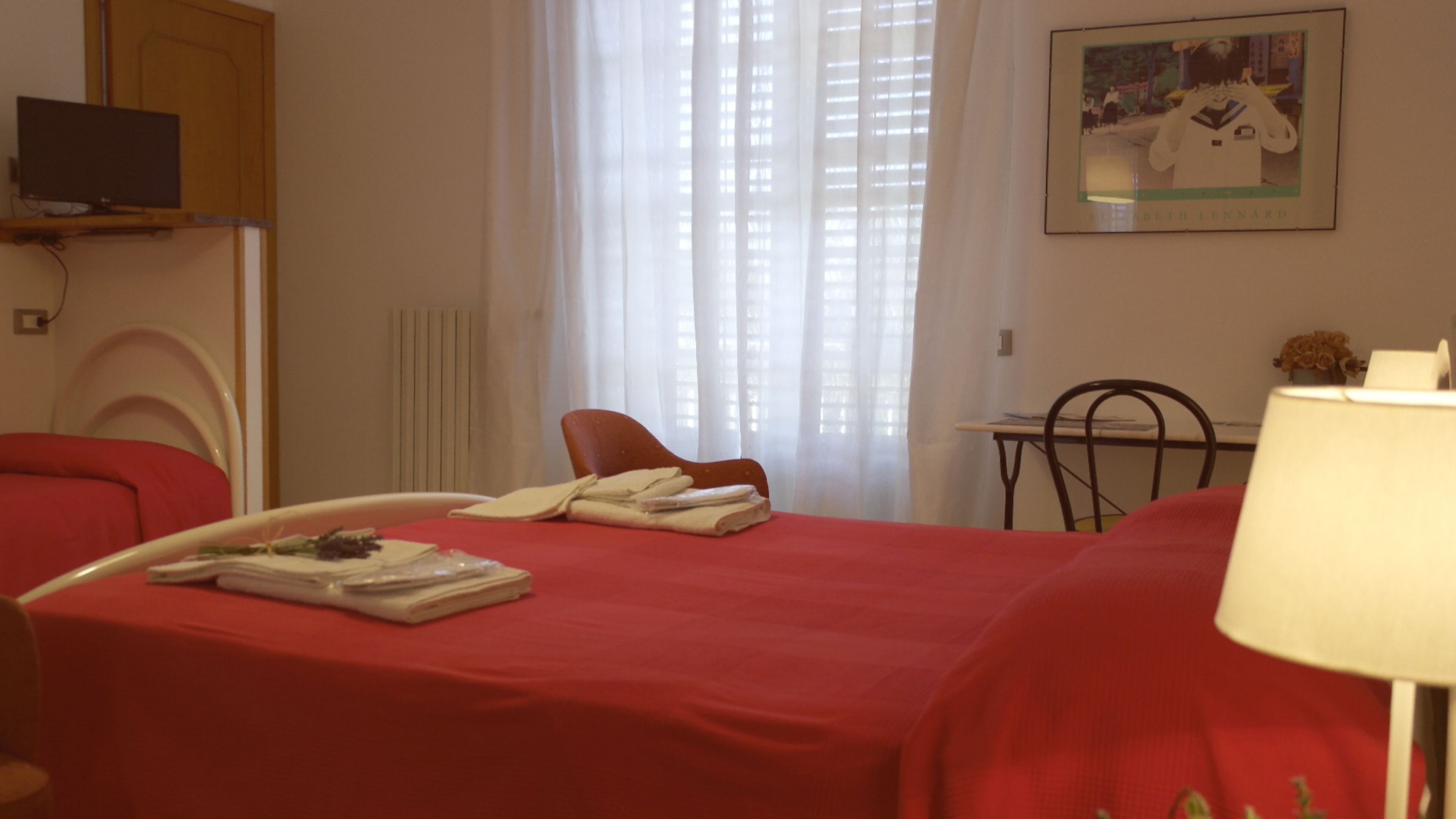 A hotel room with a red bed, a chair, a table with a lamp, a TV on a stand, and a painting on the wall.