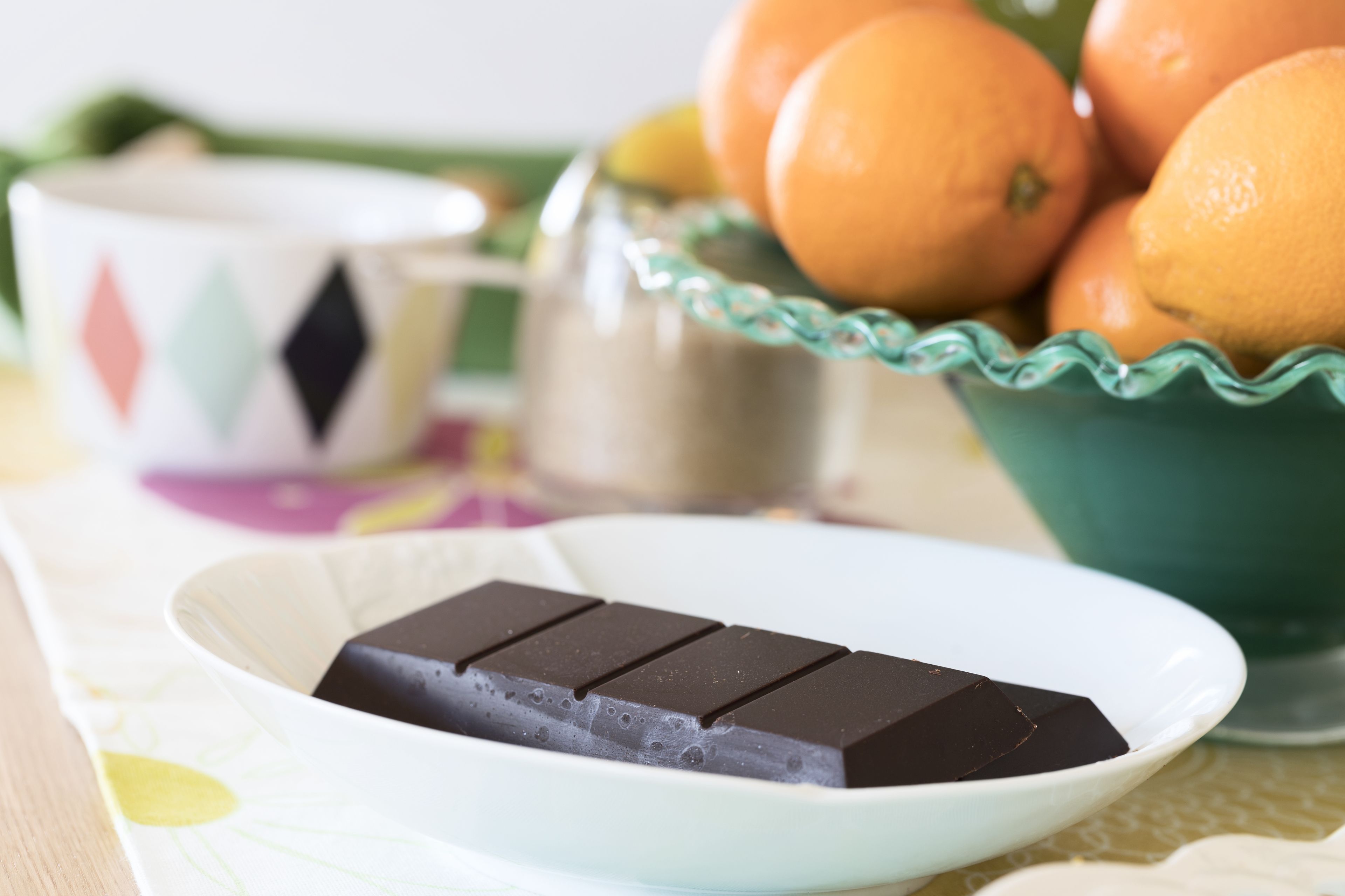 A plate with a chocolate bar, a bowl of oranges, and a teapot on a table.