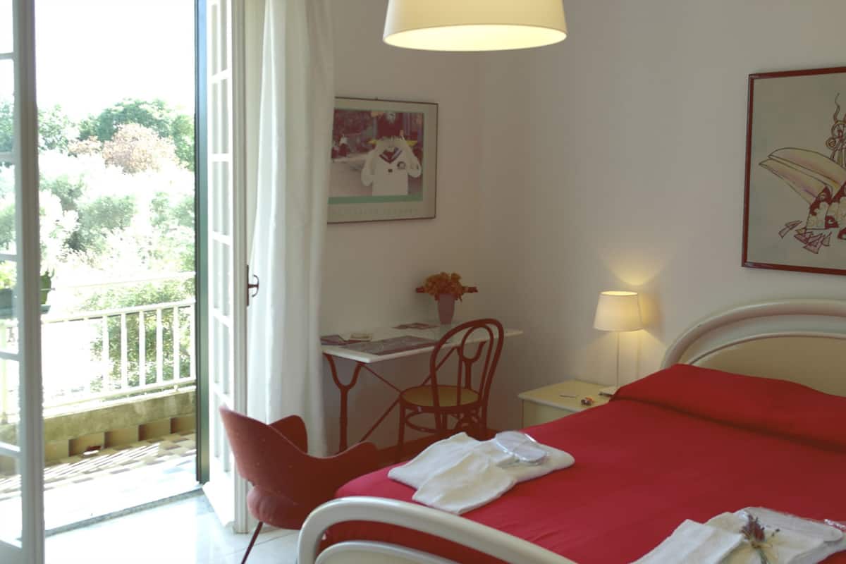 A bedroom with a bed, a desk, a chair, a lamp, and a window with a view of greenery.
