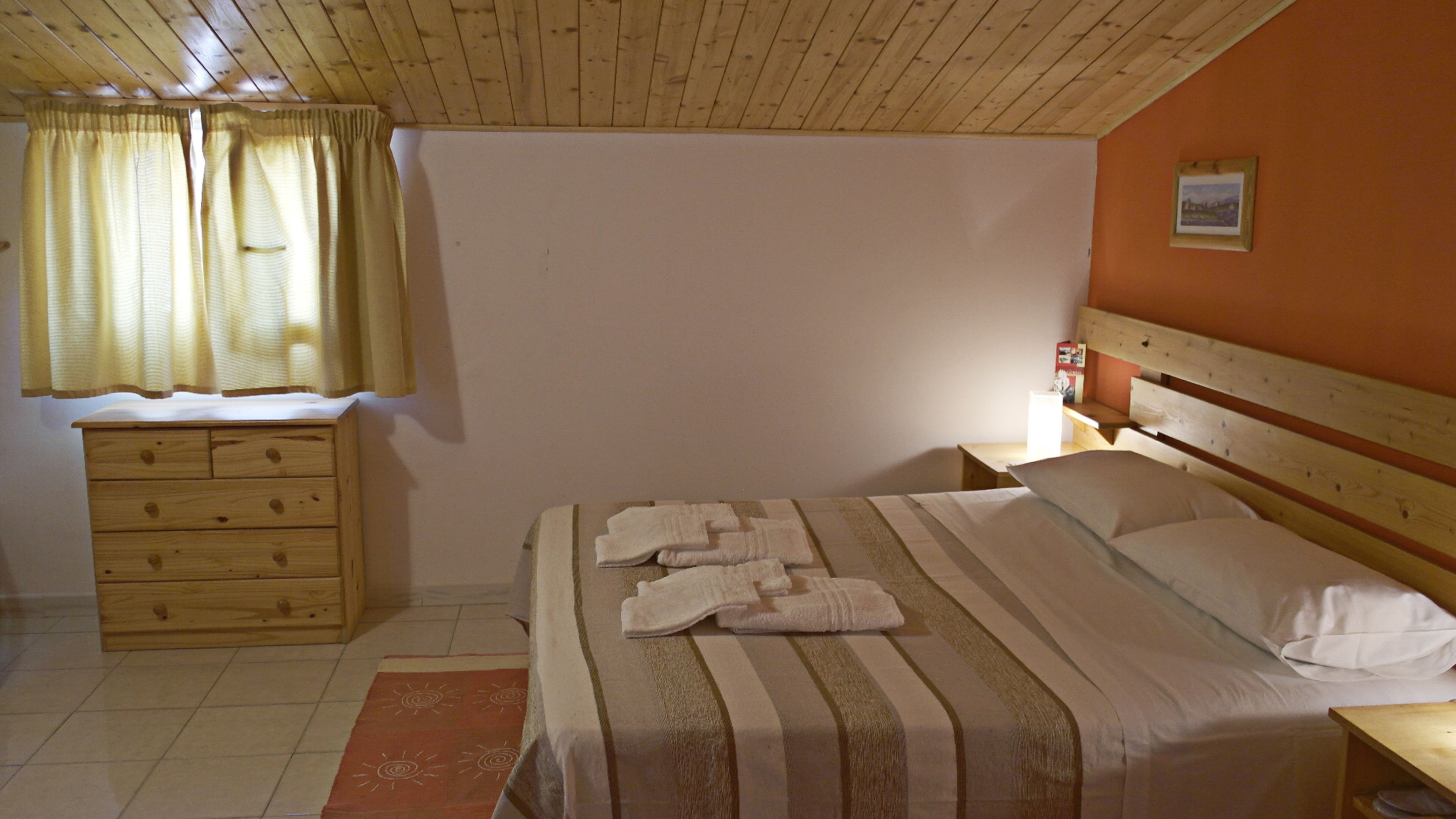 A bedroom with a wooden headboard, a bed with white linens and towels, a nightstand, a wooden dresser, and a window with curtains.