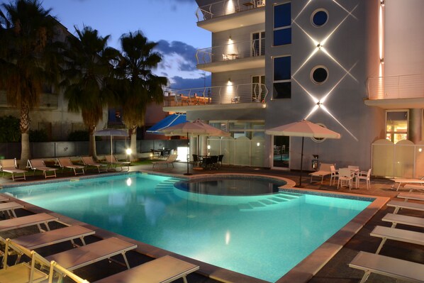 A swimming pool with lounge chairs and umbrellas, surrounded by palm trees and a building with balconies.