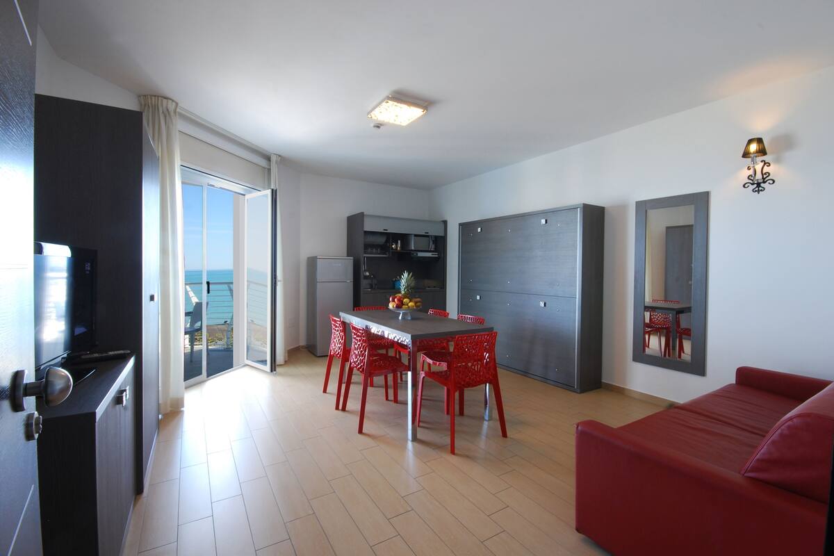 A modern living room with a red sofa, a dining area with red chairs, a kitchen with grey cabinets, and a large sliding glass door leading to a balcony.