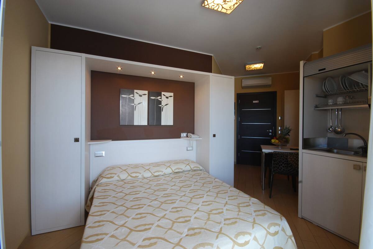 A compact hotel room with a bed, a small kitchenette, a dining area, and a bathroom.