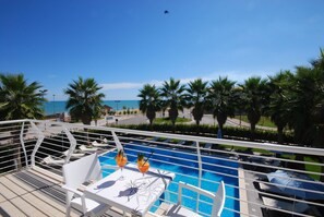 A pool area with lounge chairs and a table, surrounded by palm trees and a view of the sea.