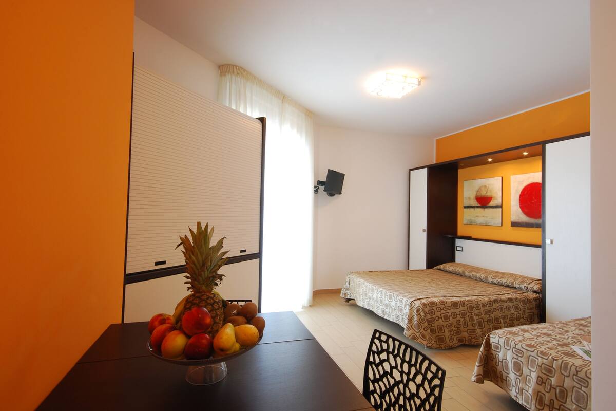 A hotel room with two beds, a dining table with fruit, and a wall-mounted TV.