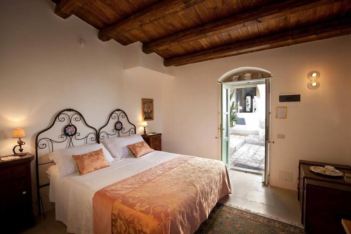 Quarto com teto de madeira, cama com colcha estampada, mesa de cabeceira com abajur e porta que dá para o exterior.