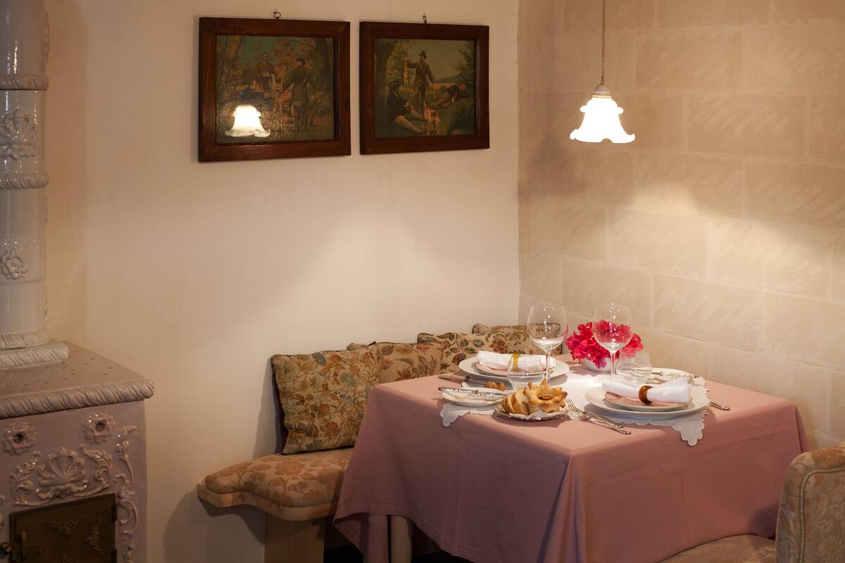 Uma área de refeições com uma mesa posta para dois, com toalha de mesa rosa, pratos, copos e um arranjo floral.