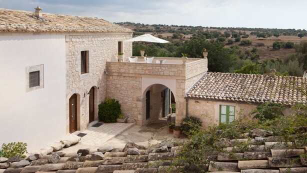 A stone building with a tiled roof, a covered outdoor seating area, and a view of the countryside. - Tenuta Cammarana (Ragusa)