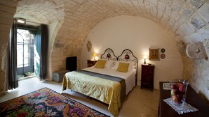 A bedroom with a stone ceiling, a bed with a green and yellow bedspread, a wooden nightstand with a lamp, and a vase of fruit on a table. - Tenuta Cammarana (Ragusa)