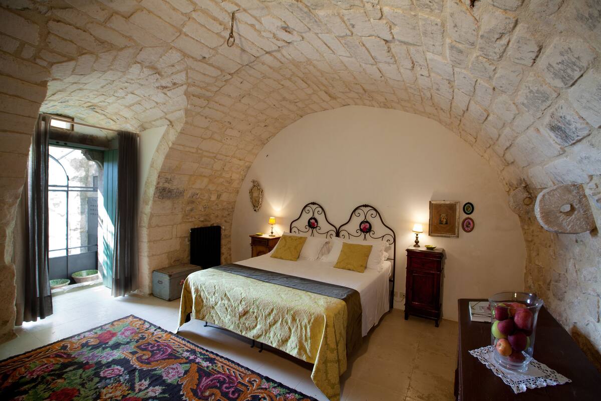 Um quarto com teto de pedra, uma cama com colcha verde e amarela, uma mesinha de cabeceira de madeira com abajur e um vaso com frutas sobre uma mesa.