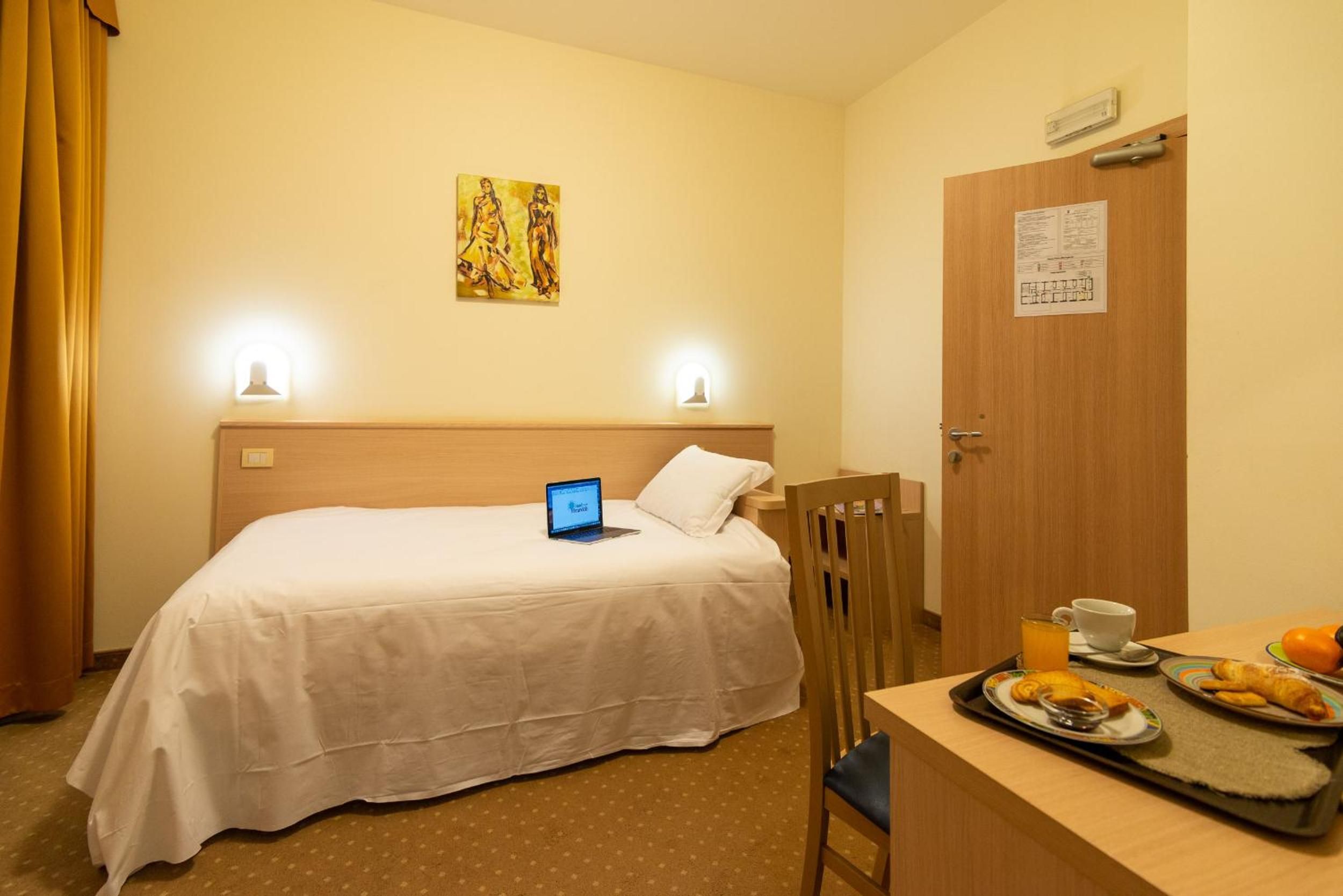 A hotel room with a bed, a laptop on the bed, a chair, a table with breakfast items, and a door with a notice.