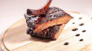 A single cooked meat item, likely a rib, on a wooden board with a few black dots around it. - Hotel Mirandola (Mirandola)