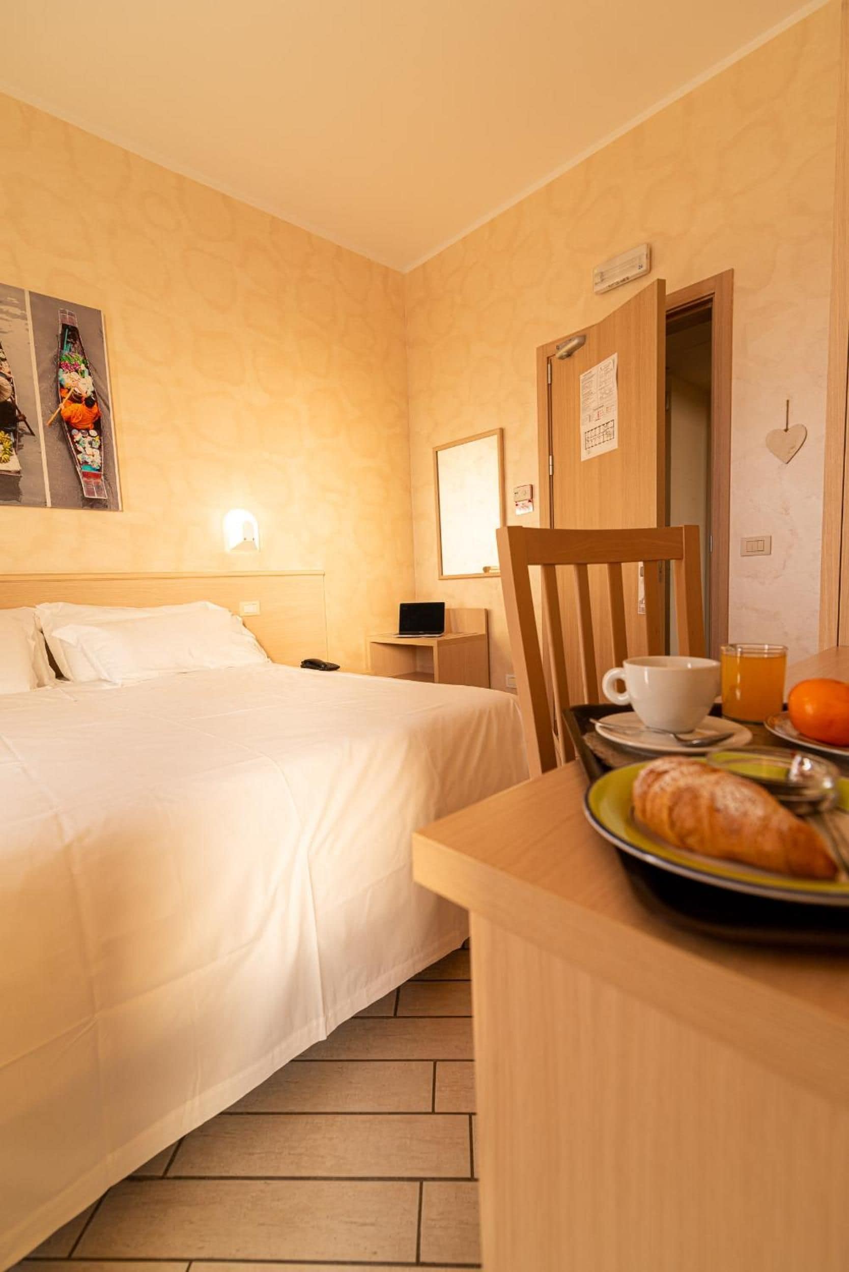 A hotel room with a bed, a bedside table, a chair, a small table with breakfast items, and a wall-mounted light.